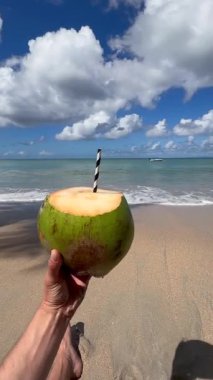 Elinde taze hindistan cevizi içeceği, arka planda mavi gökyüzü ve okyanus olan güneşli tropikal bir plajda pipetle. 