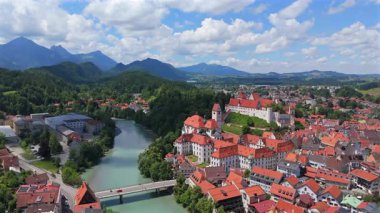Hohes Schloss şatosunu ve tarihi Fussen, Bavyera kasabasını nehir ve dağ manzaralı gösteren hava videosu 