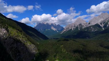 Uçsuz bucaksız, yemyeşil vadi manzarası sarp orman tepeleri ve dağılmış bulutlarla dolu mavi gökyüzünün altındaki kayalık dağ tepeleriyle çevrili.