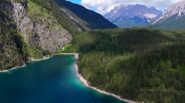 Turkuaz dağ gölü, orman kayalıkları ve yaz bulutlarının altında tepeler.
