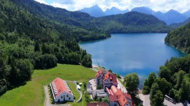 Almanya 'da Bavyera' daki Turkuaz Alpsee Gölü 'nün çevresi yemyeşil ormanlarla ve parlak bir yaz gökyüzünün altında dağ zirveleriyle çevrilidir.