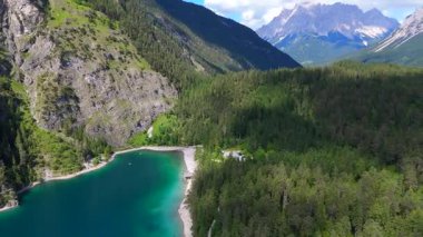 Uzak kayalık tepeleri ve turkuaz göl kıyıları olan ormanlık dağ vadisinde parçalı bulutlu bir yaz gökyüzünün altında küçük kumlu bir sahil var.