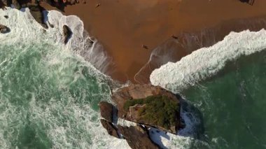 Kumlu sahilde dalgalar kırılıyor, kayalar ve insanlar kıyıda yürüyor. 