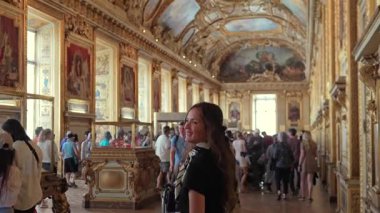 Süslü bir müze galerisinde altın tavanlar, tarihi tablolar ve 1 Haziran 2025 'te Paris' in zarif iç kısımlarını keşfeden ziyaretçilerle yürüyen genç bir kadın. Sadece editör kullanımı.