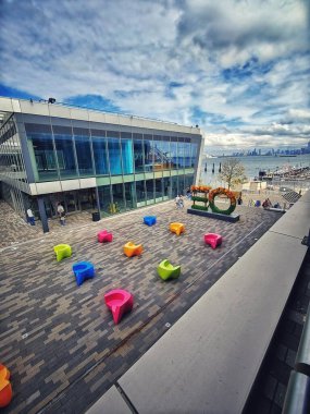 A modern urban space featuring a large plaza with colorful tiled patterns and dynamic, uniquely shaped benches. In the background, a large modern building with a glass facade reflects the sky and city views. clipart