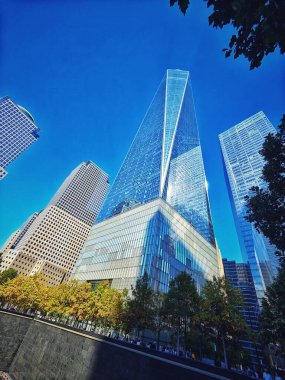 New York 'un iş bölgesinde çarpıcı bir piramit şeklindeki gökdelen mavi gökyüzünü ve etrafındaki binaları yansıtıyor. Ağaçlı yeşil bir alan kentsel çevreyle çelişiyor.