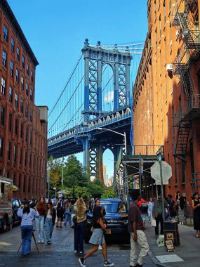 A panoramic view of New York with the Brooklyn Bridge crossing the East River, its elegant arches standing out against the vibrant sky and calm waters. The skyline of Manhattan, including One World Trade Center, towers in the background. clipart