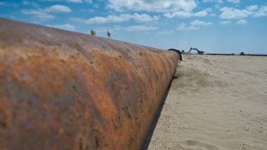 Büyük paslı bir endüstriyel boru, deniz kenarındaki kumlu bir plaj boyunca uzanır ve mavi gökyüzünün altında inşaat makineleri görünür.. 