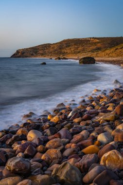 Hallett Cove Sahili 'nde gün batımında sakin bir sahil manzarası. Yavaş bir kepenk hızıyla bulanıklaşan hafif dalgalar renkli, yuvarlak kayalardan oluşan bir plajın üzerinde süzülür.. 