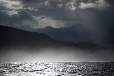 Fırtınalı bir deniz manzarasının büyüleyici bir fotoğrafı sisli dalgalar ve dramatik bulutlarla dolu bir gökyüzünün altındaki karanlık dağlık kıyı şeridi.