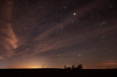 Litvanya 'daki siluetli bir peyzaj ve nirengi kulesinin üzerinde yıldızlarla dolu göz kamaştırıcı bir gece gökyüzünün geniş açılı görüntüsü. Görünür takımyıldızları ve ufuktaki sıcak ışık kirliliği olay yerine derinlik ve atmosfer ekler..
