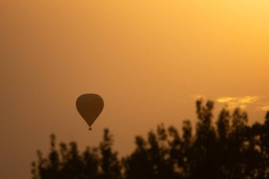 Sıcak hava balonu zarif bir şekilde gün batımının turuncu parıltısında süzülür, parlak altın gökyüzüne karşı siluetlenir. Ağaç tepelerinin koyu çizgisi kontrastı güçlendirir ve huzur sahnesine derinlik katar. Bu görüntü özgürlük, huzur ve... 