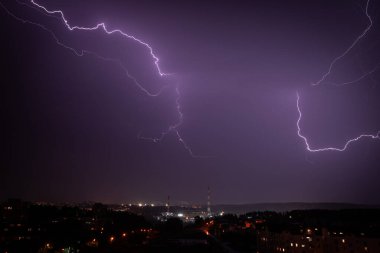 Uzak bir şehir manzarasının üzerinde gece gökyüzünde çarpıcı şimşekler çakar ve aşağıdaki binaların üzerine ürkütücü bir menekşe parıltısı saçar. Görüntü, bir fırtınanın ham gücünü şehir perspektifinden yakalıyor..