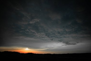 Güneş ufkun altına batarken gökyüzünde hareket eden büyüleyici bir memeli bulutu alanı. Ağır, dokulu oluşumlar, günün son altın ışığında yıkanan sakin bir manzaranın üzerinde beliriyor. Fırtınalı gerilim ve günbatımı arasındaki çarpıcı tezat.