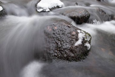Yumuşak, koyu renkli kayalar, kısmen ince kar ve buz tabakalarıyla kaplı, yumuşak bir kış nehrinde otururlar. Su onların etrafında yumuşak, ipeksi çizgiler halinde akar, hareket ve durgunlukla dolu meditasyon kompozisyonu yaratır..