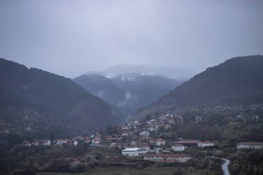 Sessiz bir köy, orman kaplı tepelerin eteklerinde kısmen kayan dağ sisi ile örtülü duruyor. Bulutlar tepeleri yumuşak katmanlarla örterken ortam sakin ama gizemli..