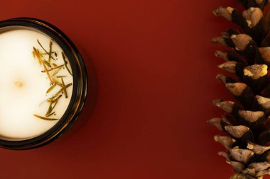 A top down view of a decorative aromatic candle in a glass jar, accompanied by a natural pinecone, all set against a bold red background. The minimalist composition is ideal for holiday themes clipart