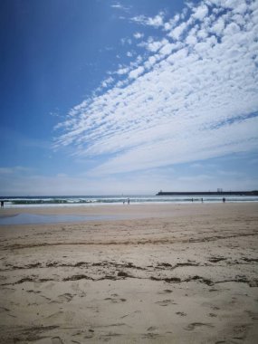 Parlak mavi gökyüzünün altında dağınık bulutlu canlı bir plaj sahnesi. Yumuşak dalgalar kumlu sahile ulaşır ve birkaç insanın günün tadını çıkardığı uzaklardan görülebilir. Seyahat ve turizm için mükemmel.