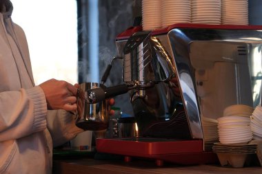 Modern bir kahve makinesi kullanarak, bir baristanın metalik sürahide süt buharlaştırdığı samimi bir kahve dükkanı sahnesi. Açık renk kapüşonlu giyinen barista odaklanarak çalışır.