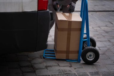 A delivery worker loading a large cardboard box onto a blue hand truck beside a van. Perfect for logistics, shipping, delivery, and transportation-related content clipart