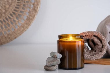 Kehribar mum kavanozu ve parlayan bir alev içeren rahatlatıcı bir kompozisyon, bej gölgelere sarılmış havluların yanına yerleştirildi. Sahne, hasır elementlerin oluşturduğu doğal bir arkaplan üzerine kuruludur.