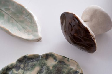 Beyaz bir yüzeyde seramik dekorasyonların yakın çekimi. Parlak yeşil bir yaprak tabağı, desenli bir mantar heykeli ve botanik izleri olan soyut yeşil bir tabağı içeriyor. Şey...