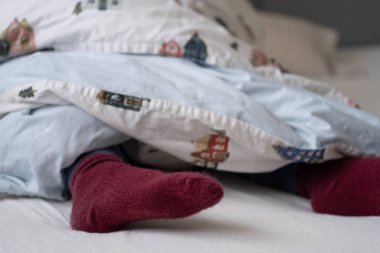 A cozy close up of maroon socks peeking out from under a colorful duvet with a whimsical house pattern. The setting exudes warmth and comfort, making it suitable for advertisements for cozy bedding clipart