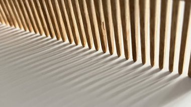 Beyaz bir yüzeyde duran, doğal ışığın yansıttığı dramatik gölgeleri olan ahşap bir tarağın görüntüsünü kapat. Bileşim, tarağın dişlerinin dokusunu ve desenini vurgular, soyut ve soyut bir şekil yaratır.