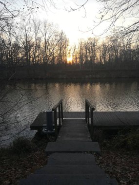 Huzurlu bir ahşap iskele sakin bir göl üzerinde uzanır, arka planda altın gün batımı parıltısı vardır. Nehir kıyısındaki çıplak ağaçlar sakin kompozisyonu tamamlıyor. Seyahat dergileri için uygun, sakinleştirici.