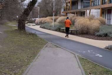 A solitary runner is captured from the back jogging along a well-paved path lined with ornamental grasses and modern apartment buildings in the background. The vibrant orange jacket of the runner clipart