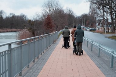 Bulutlu bir kış gününde, üç yetişkinin puslu bir kış günü bebek arabalarıyla asfalt bir nehir kenarında yürüdükleri huzurlu bir manzara. Soldaki nehir kısmen donmuş ve yaprakları olmayan ağaçlar her iki tarafta da sıralanmış.