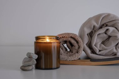 Kehribar rengi cam bir mum kavanozu, Mocha Mousse tonlarında havluların yanına yerleştirilmiş, ahşap bir ayak fırçasının üzerinde dinleniyor. Sahne, istiflenmiş doğal taşlarla tamamlanmıştır.