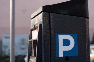 A closeup view of a modern parking meter with a blue P sign on its side. The surface is covered in water droplets, emphasizing its outdoor location. The blurred background features urban elements. The clipart