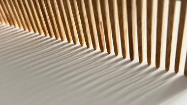 Beyaz bir yüzeyde duran, doğal ışığın yansıttığı dramatik gölgeleri olan ahşap bir tarağın görüntüsünü kapat. Bileşim, tarağın dişlerinin dokusunu ve desenini vurgular, soyut ve soyut bir şekil yaratır.