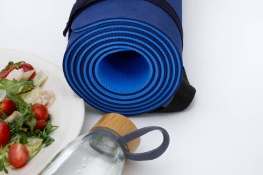 A healthy lifestyle composition featuring a blue yoga mat, a bamboo-lid water bottle, and a plate of fresh salad with cherry tomatoes and lettuce. Photographed on a white background, ideal for clipart