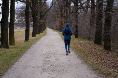 Serin bir kış gününde, ağaçlarla kaplı sakin bir patikada koşan mavi atletizm kıyafetleriyle tek bir koşucu. Uzun ağaçlarla çevrili uzun yol, sakin ve meditasyon yapan bir atmosfer yaratır. Bu resim