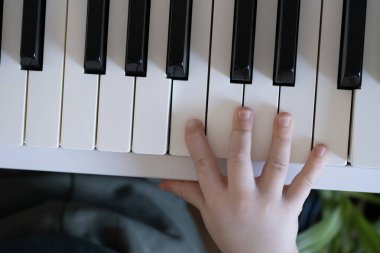 Genç bir çocuğun sağ kolu beyaz bir piyano klavyesinde dinleniyor. O an müziğin nazik ve doğal keşfini dokunarak yakalar. Müzik eğitimi promosyonları ve çocukluk dönemi için uygun