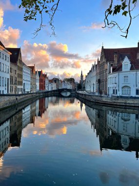 Belçika 'nın Bruges kentindeki bir kanalın manzarası. Gün batımında sakin sulara yansıyan tarihi binalar. Gökyüzü yumuşak pastel renklerle boyanır. Huzurlu ve resimsel bir atmosfer yaratır.
