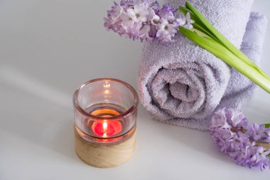 Sarılmış lavanta havlusu, taze sümbül çiçekleri ve minimalist beyaz bir arkaplanda yanan bir mumun yer aldığı kaplıca esaslı bir düzenleme. Kompozisyon ışıldıyor.