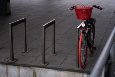 A full view of a black bicycle with red tires, a red basket, and a pink lock, parked in a designated bike rack area. The composition highlights urban cycling and eco-friendly transportation. Perfect clipart