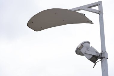 Bulutlu beyaz gökyüzüne karşı koruyucu metal kaplı modern bir sokak lambasının yakın çekimi. Tasarım, çağdaş kentsel altyapı ve açık hava altyapısını vurguluyor ve düzgün ve işlevsel görünüyor.