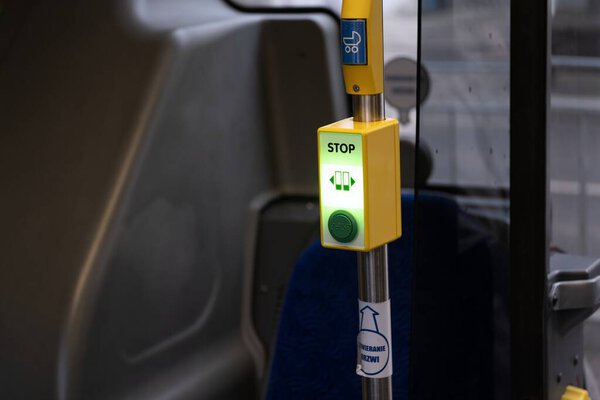 Крупный план кнопки остановки внутри общественного автобуса с зеленым индикатором "Стоп", указывающим на активированный запрос. Кнопка крепится к желтому столбу, а синее сиденье - к синему.