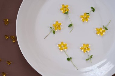 D vitamini softgel kapsüllerinin beyaz bir tabakta narin çiçek şekilleri oluşturması için yaratıcı bir düzenleme. Kapsüllerin altın rengi, temiz, minimalist arkaplanla çelişiyor.