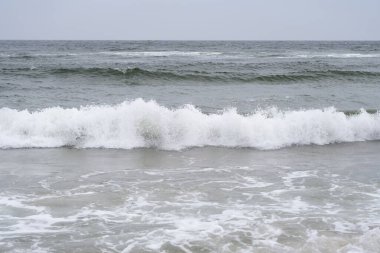 A dynamic ocean wave crashes onto the shore, creating a dramatic display of white foam and swirling water. The energy of the sea is captured in this high-resolution image, with deep blue and green