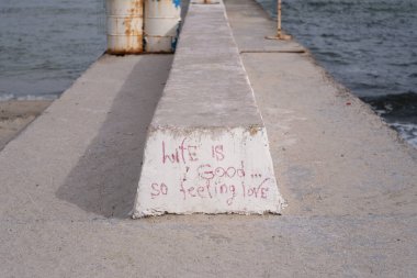 Yıpranmış beton bir iskeleye yakın çekim. El yazısıyla yazılmış grafiti ile hayat güzel. Yani aşkı hisset. Deniz dalgaları arka planda çarpar ve insan ifadesi ile doğal ifade arasında kontrastlar oluşturur.