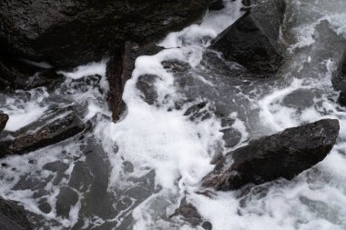 Beyaz deniz köpüğüyle çevrili koyu renkli kayalara yakın çekim. Keskin taş kenarlar ve akan su arasındaki zıtlık dinamik ve dokusal bir görüntü yaratıyor, arka planlar, bloglar ve tasarım şablonları için uygun.
