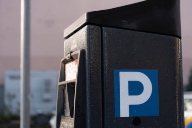 Yan tarafında mavi P işareti olan modern bir parkmetrenin yakın görüntüsü. Yüzey su damlacıklarıyla kaplı, açık hava konumunu vurguluyor. Bulanık arka planda kentsel unsurlar var. Şey...