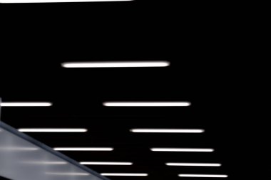 Karanlık bir iç mekanda parlayan bir dizi modern dikdörtgen tavan lambasının soyut görüntüsü. Geometrik hizalama ve gölge ve ışık arasındaki zıtlık minimalist ve gelecekçi bir atmosfer yaratır.