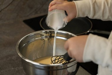 Küçük bir el çırpıcı ve ölçü kabını tutar ve krep hamuruna süt döker. Kadehten ince bir süt akıntısı akar. O, çok rahat ve temiz bir mutfakta karanlık yumuşak tonlarda ışık altında yemek pişiriyor. Ailesinin mutfağında iyi bir yardımcı.. 