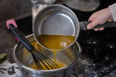 Küçük bir çocuğun eli dikkatlice eritilmiş malzemeleri bir tencereden çırpma kabıyla karıştırma kabına döküyor ve pişirme işlemine devam ediyor. Bu an, yemek tarifleri, çocuk dostu yemekler ve duyular için mükemmel olan erken mutfak müdahalesini yakalar.